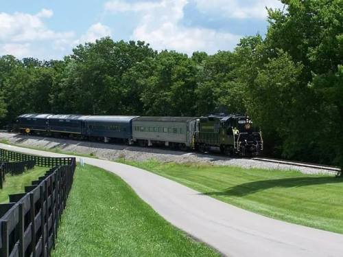 A vintage train travels along a curved track surrounded by lush green trees and a paved path.