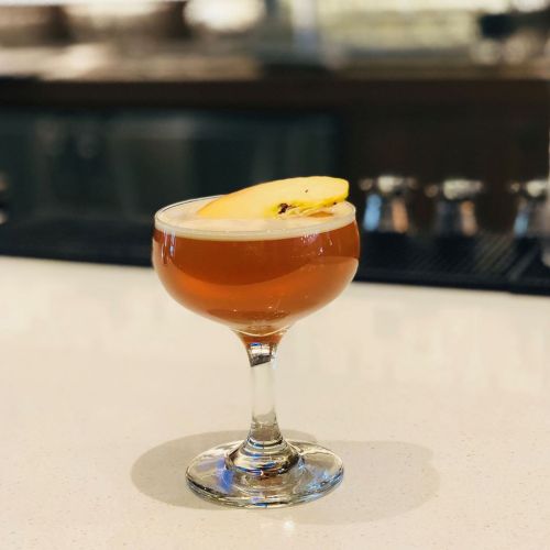 A cocktail in a coupe glass, garnished with a slice of apple, sits on a bar counter.