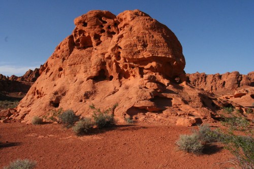A large, reddish rock formation surrounded by sparse vegetation under a clear blue sky.
