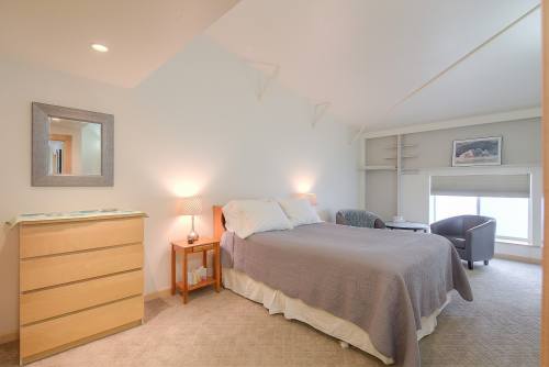 Cozy bedroom with a queen bed, dresser, armchair, and soft lighting, featuring a large window and neutral decor.