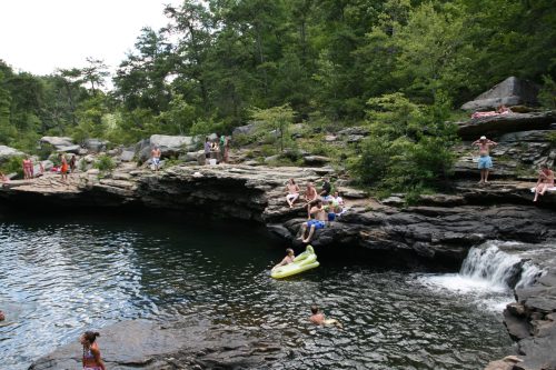 A natural swimming area with people lounging on rocks, swimming, and enjoying the water surrounded by trees.