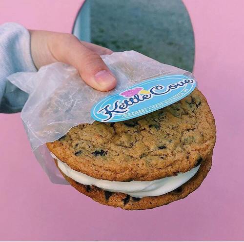A hand holds a cookie sandwich with ice cream, wrapped in paper, against a pink background. Kettle Cove logo visible.