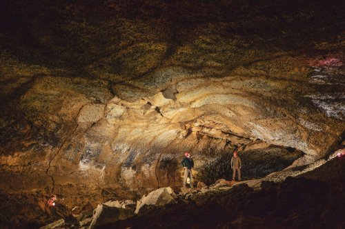 Two explorers stand on rocky terrain inside a cave, illuminated by their headlamps, showcasing textured stone walls above.