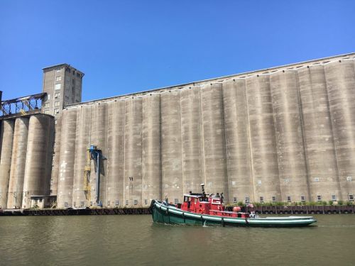 A red tugboat navigates a river near large concrete silos under a clear blue sky.