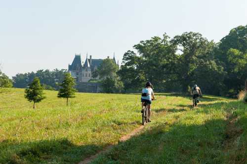 Two cyclists ride on a grassy path with a castle in the background under a clear blue sky.