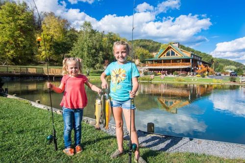 Two young girls hold fishing rods and caught fish by a pond, with a lodge and trees in the background under a blue sky.