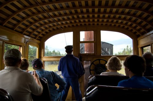 Interior view of a vintage train, with passengers seated and a conductor at the front, overlooking scenic landscapes.