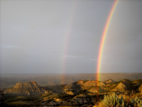 A scenic view of rolling hills under a cloudy sky, featuring two vibrant rainbows arching over the landscape.