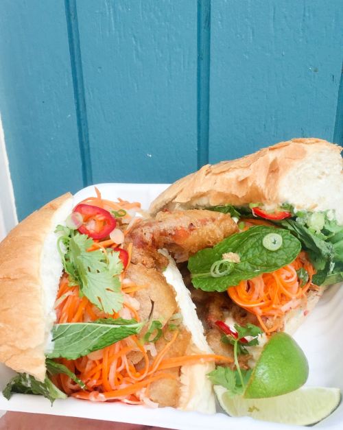 A close-up of a Vietnamese banh mi sandwich filled with fried ingredients, fresh herbs, and colorful vegetables, served with lime.