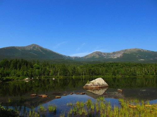 A serene landscape featuring a calm lake reflecting mountains and lush greenery under a clear blue sky.