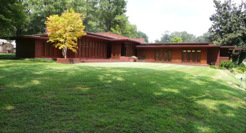 A modern, single-story wooden house surrounded by lush green grass and trees on a sunny day.