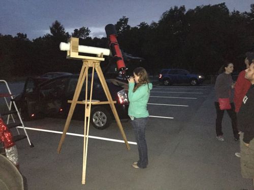 A person looks through a telescope set up in a parking lot at dusk, with others nearby and cars in the background.