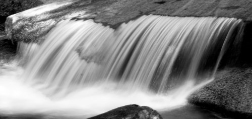 A black and white image of a gentle waterfall cascading over smooth rocks.