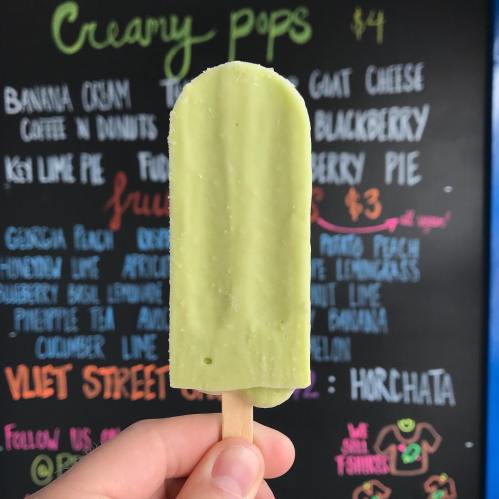 A hand holding a green popsicle in front of a colorful menu board featuring various flavors and prices.