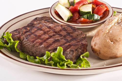 A grilled steak on a plate with lettuce, a baked potato, and a side of mixed vegetables in sauce.