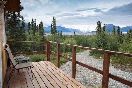 A wooden deck with two chairs overlooks a scenic view of mountains and greenery under a cloudy sky.