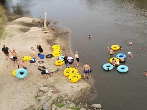 A group of people enjoying a river, some in the water with inner tubes, others on the shore with yellow and blue floats.