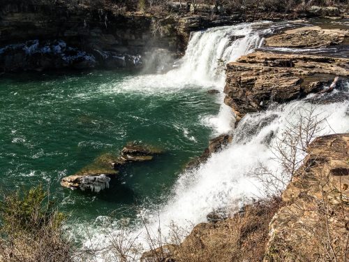 A scenic view of a waterfall cascading over rocky cliffs into a turquoise river, surrounded by trees and natural landscape.