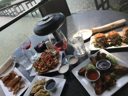 A table with various dishes including nachos, sliders, wings, and drinks, overlooking a scenic waterfront view.