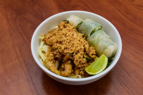 A bowl of noodles topped with chicken, crushed peanuts, and fresh spring rolls, garnished with a lime wedge.
