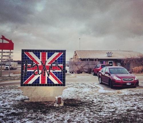A sign featuring a British flag design outside a restaurant, with a car parked nearby and a cloudy sky above.