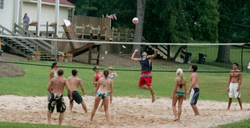 A group of people playing beach volleyball on a sandy court surrounded by trees and a building in the background.