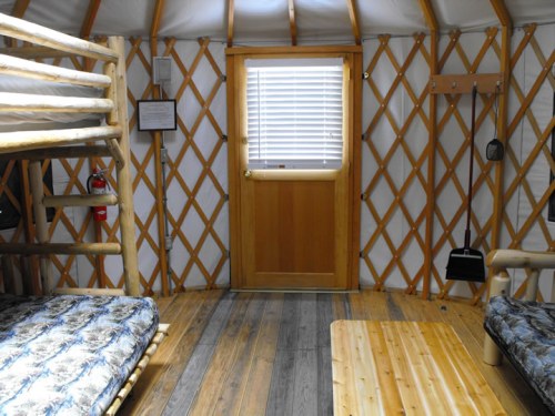Interior of a yurt featuring bunk beds, a wooden table, and a door with a window, all surrounded by wooden lattice walls.