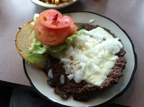 A plate with a hamburger patty topped with onions, lettuce, tomato, and mayonnaise, served with a bun on the side.