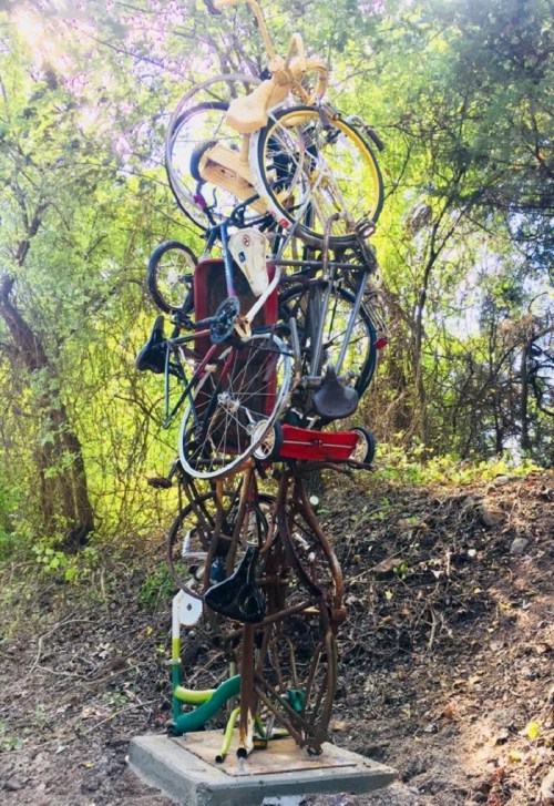 A sculpture made of stacked bicycles and bike parts, surrounded by greenery in a natural setting.