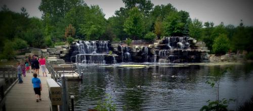 A serene park scene featuring a waterfall, lush greenery, and a wooden walkway with people enjoying the view.