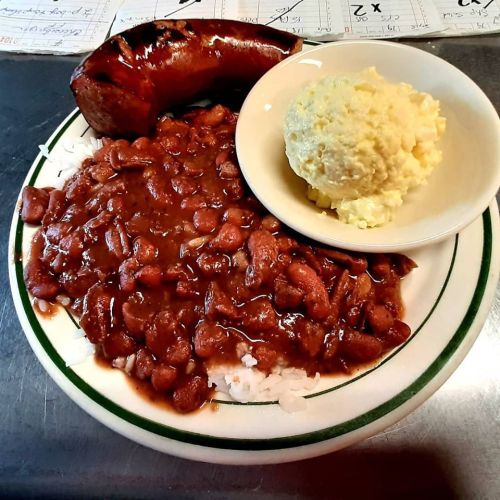 A plate of rice topped with red beans, served with a sausage and a side of creamy potato salad.