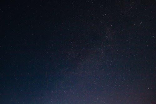 A clear night sky filled with countless stars, showcasing the Milky Way and a faint meteor streak.