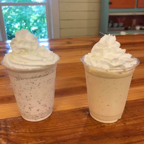 Two cups of milkshakes topped with whipped cream, one with chocolate bits and the other plain vanilla, on a wooden table.