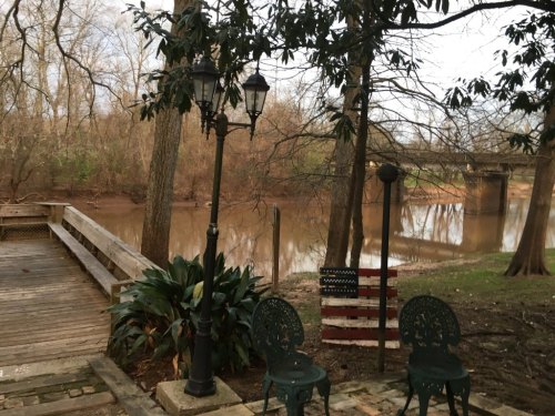 A peaceful riverside scene with a wooden deck, two green chairs, and a decorative American flag near a calm river.