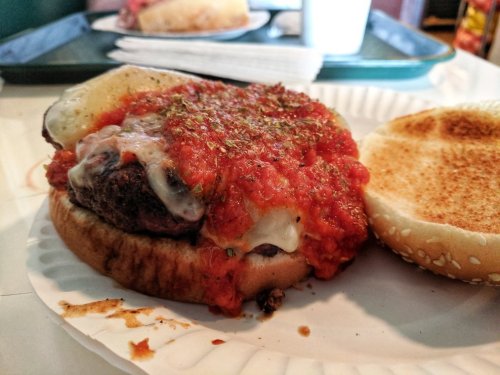A close-up of a burger with melted cheese, marinara sauce, and herbs on a sesame seed bun, served on a paper plate.