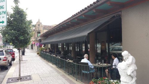 Outdoor dining area of a restaurant with awnings, tables, and a statue, located on a city street.