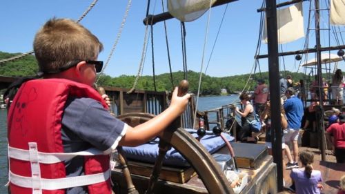 A child in a life jacket steers a pirate ship on a sunny day, surrounded by people enjoying the boat ride.