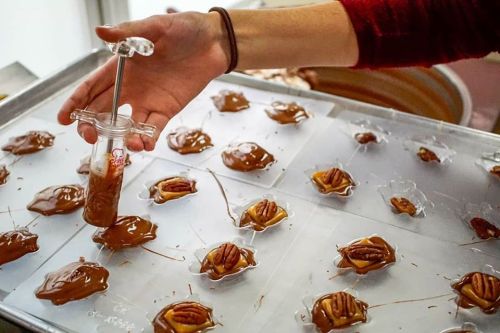 A hand holds a syringe over a tray of chocolate-covered pecans on parchment paper.