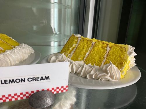A slice of lemon cream cake with yellow layers and white frosting, displayed on a plate next to a sign.