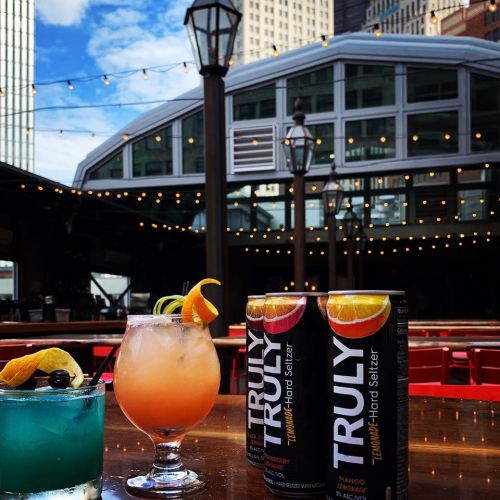 Two colorful cocktails and cans of Truly Hard Seltzer on a table, with a cityscape and string lights in the background.