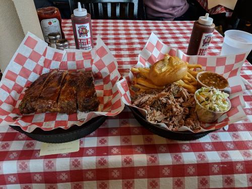 Two plates of barbecue: one with ribs and fries, the other with pulled pork, coleslaw, beans, and a bun, on a checkered tablecloth.