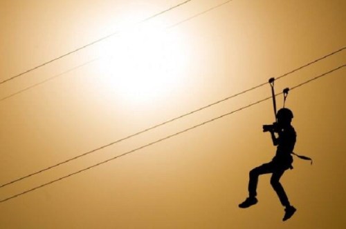 A silhouette of a person zip-lining against a bright sunset, holding a camera.