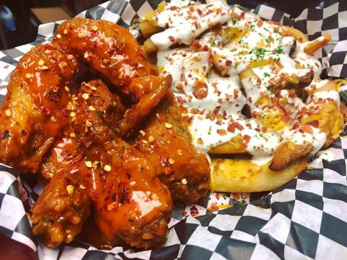 A plate of spicy chicken wings and loaded fries topped with cheese, bacon, and sour cream, served in a checkered basket.
