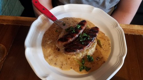 A plate of grilled sausages on creamy mashed potatoes, garnished with parsley and served with a rich sauce.