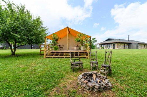A cozy tent with a wooden deck, surrounded by greenery and a fire pit with stone seating in a serene outdoor setting.