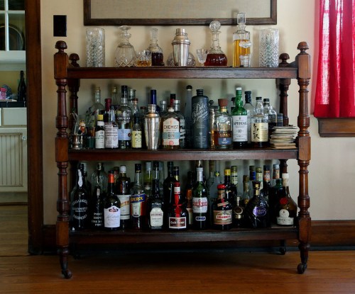 A wooden bar cart with multiple shelves displaying various bottles of liquor and glassware in a cozy room.