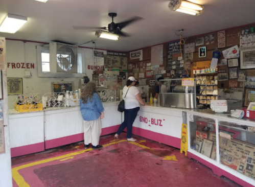 A colorful ice cream shop interior with two customers at the counter, surrounded by vintage signs and a fan overhead.