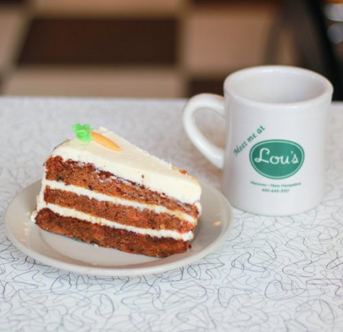 A slice of carrot cake with cream cheese frosting on a plate, next to a coffee mug that says "Meet me at Lou's."
