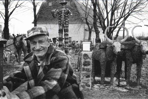 A smiling man in a checkered coat stands in a garden filled with unique stone sculptures of animals and figures.