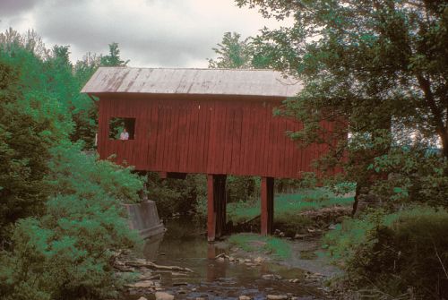 A red covered bridge stands on stilts over a small stream, surrounded by lush greenery and trees.